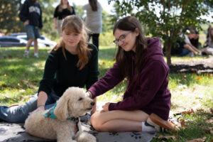 Two girls sit on a blanket in a park and pet a fluffy white poodle wearing a turquoise bandana.
