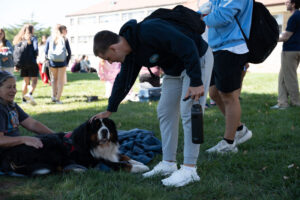 Man bending to pet a large Bernese Mountain Dog lying on a blanket on a grassy campus lawn, with a woman sitting nearby and students standing in the background.