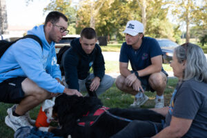 Group of four people kneeling on grass around a large black and tan dog wearing a red harness, outside on a sunny day in a park setting.