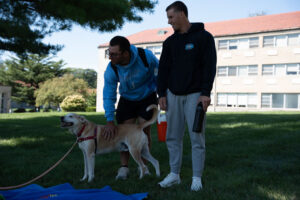 Two men stand on a grassy campus lawn with a tan dog on a leash; one man pats the dog while the other holds a water bottle nearby.