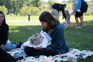 Woman in a denim jacket sits on a blanket in a park, cradling a small fluffy dog wrapped in a towel.
