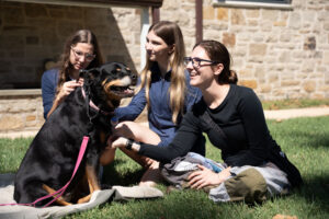 Three women sit on a blanket on the grass outside a stone building, smiling as they pet a black and tan dog on a pink leash.