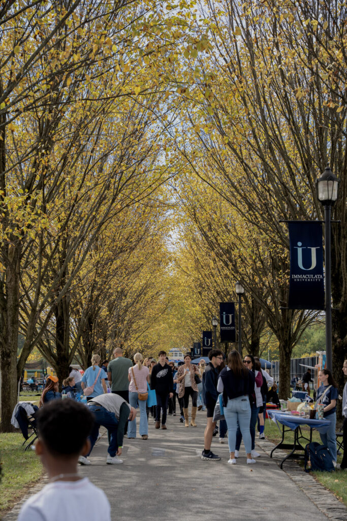 Group of students walking and chatting along a tree-lined campus path during an outdoor event, banners on lampposts visible under yellow autumn leaves.