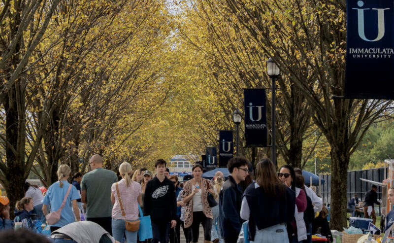 Group of students walking and chatting along a tree-lined campus path during an outdoor event, banners on lampposts visible under yellow autumn leaves.
