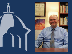 Composite graphic with a navy blue background featuring a light blue silhouette of a domed building topped with a cupola and statue figure on the left, and a photo of David Martinson, a smiling man with a short gray beard, wearing a light blue shirt and patterned tie—seated at a desk in an office on the right.