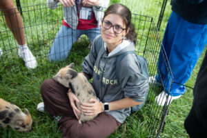 Young woman with glasses sits on grass inside a small pen, cradling a piglet while others look on at an outdoor animal event nearby.