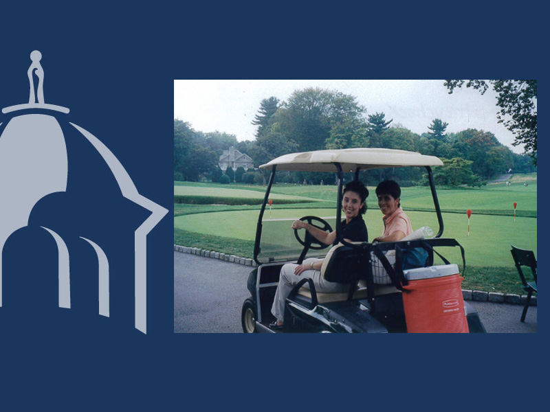 Photo of two women riding in a golf cart on a golf course path, displayed on a dark navy blue background with a light gray silhouette logo of a building topped with a small statue figure in the lower left corner. In the photo, one woman drives the cart wearing a black polo shirt and light pants, smiling at the camera; the other, in a coral shirt, sits beside her. The cart carries an orange cooler and golf bag. Behind them is a golf course fairway with flagged holes, trees, and a large house in the distance.