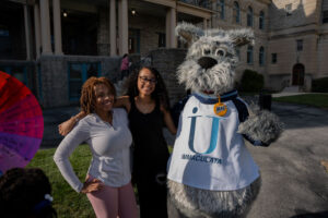 Two smiling women pose with a gray furry mascot wearing an Immaculata shirt outside a stone-campus building.