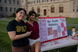 Two young women stand beside a campus information board titled 'Her Health Initiative' at an outdoor table with pamphlets and snacks nearby.
