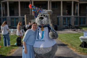 Woman in a light-blue dress poses with a gray dog mascot wearing a colorful party hat and a white IMMACULATA jersey at an outdoor campus event.