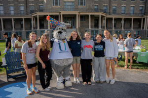Group of students posing with a gray dog mascot wearing a colorful party hat outdoors in front of a brick building during a campus event.