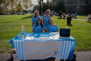 Four volunteers in blue shirts behind a booth for Immaculata University Dance Company at an outdoor campus event.