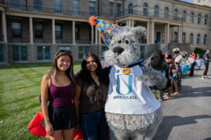 Two smiling students posing with a gray dog mascot wearing a party hat and an Immaculata jersey outside a campus building at an outdoor event.