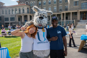 Three people pose with a gray dog mascot in an Immaculata University jersey on a sunny campus quad, smiling for the camera.