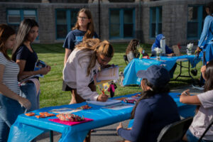 Group of people at a sunny outdoor event around a table with a blue tablecloth; a woman signs a sheet while others watch.