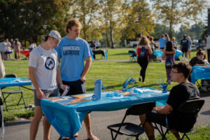 Two young men stand at a blue-clothed table at an outdoor lacrosse event, a volunteer seated nearby and others in the park behind them.