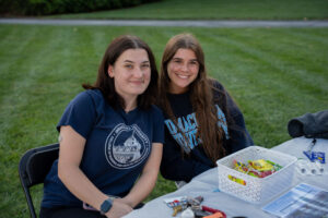 Two young women sit at a table on a grassy lawn, smiling at the camera, wearing dark blue university shirts with logos.