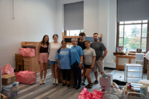 Group of eight people posing together in a room with shelves, boxes, and pink gift bags—likely volunteers organizing supplies.