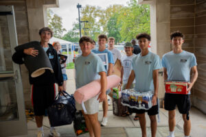 Group of volunteers carrying donation items outside a building, smiling at the camera, preparing to load supplies like boxes and water packs.