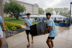 Two young men in light blue shirts carry a large black box along a rainy campus walkway.