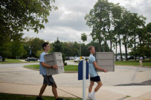Two people walk side by side outdoors, each carrying a gray plastic filing cabinet toward a parking lot.