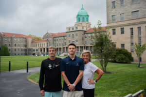 Three people pose for a photo on a university campus lawn, with historic stone buildings and a turquoise-domed tower in the background.