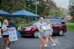 Two young volunteers in light blue shirts unloading supplies from a maroon van under blue canopy tents at an outdoor event.