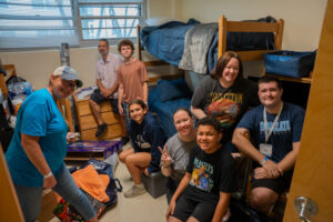 Group of volunteers smiling in a dorm room with two bunk beds and assorted supplies nearby,