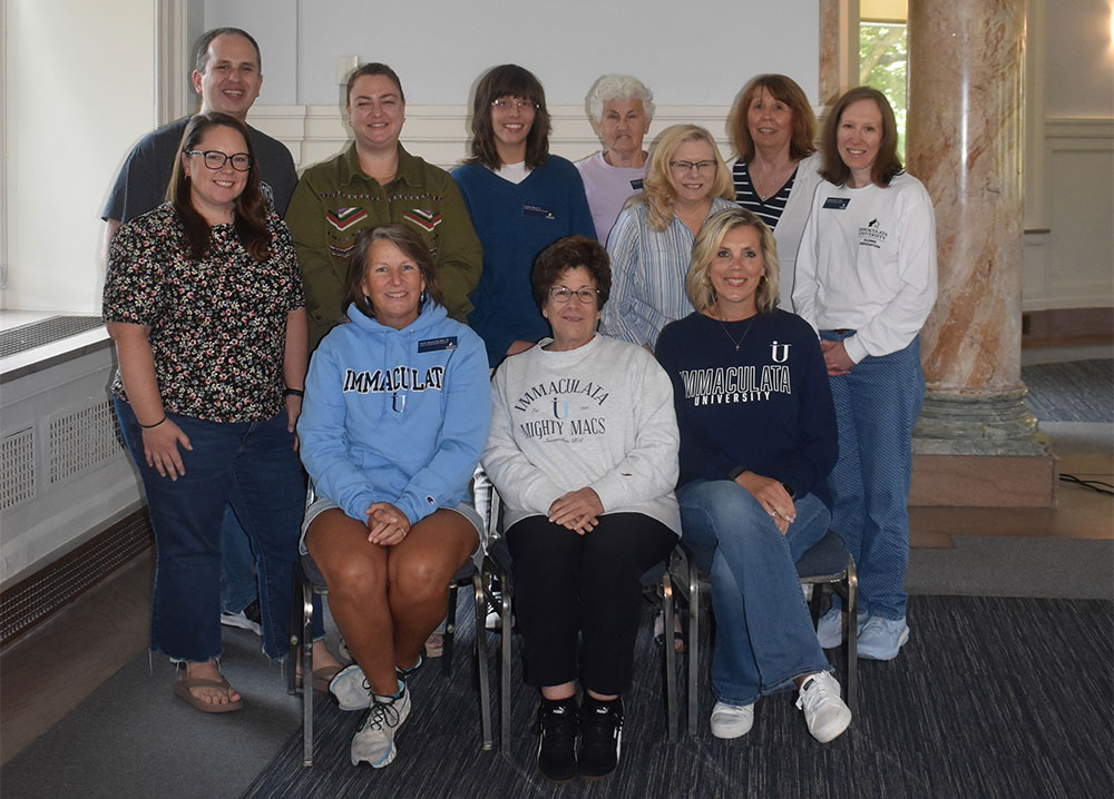aabog - Immaculata University Smiling group of twelve people posing in a campus hallway, several wearing Immaculata University sweatshirts.