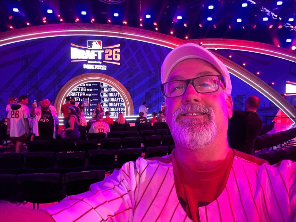 Mark McFadden, Ed.D., takes a selfie at the MLB Draft during MLB All-Star Week. Behind him, attendees sit and stand near a stage displaying “MLB Draft 26” graphics. Pink and purple lighting illuminates the event space.