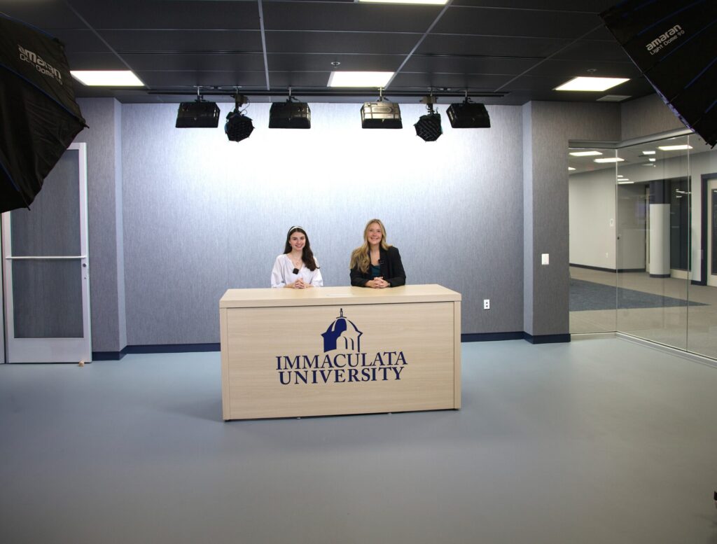 Two women sit at a wooden desk with the Immaculata University logo, in a studio with overhead lights above them.