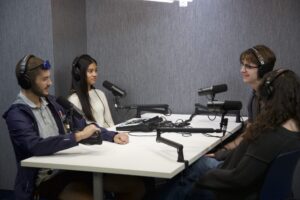 Four people wearing over-ear headphones sit around a white table in a small studio with gray textured walls. Each seat has a black microphone on an adjustable boom arm, and a small audio mixing console sits in the middle of the table. The young man at left is speaking, and the others are looking toward him or each other.