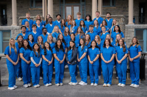 Large group of students in blue scrubs posing on the steps of a stone building outside the Back Campus entrance to Villa Maria Hall.