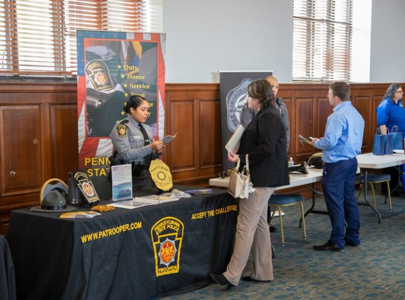 A Pennsylvania State Police representative speaks with attendees at a career fair booth displaying recruitment materials, uniforms and trooper insignia.